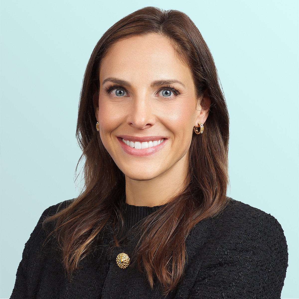 A woman with long brown hair wearing a black textured top and gold hoop earrings smiles at the camera against a light blue background.