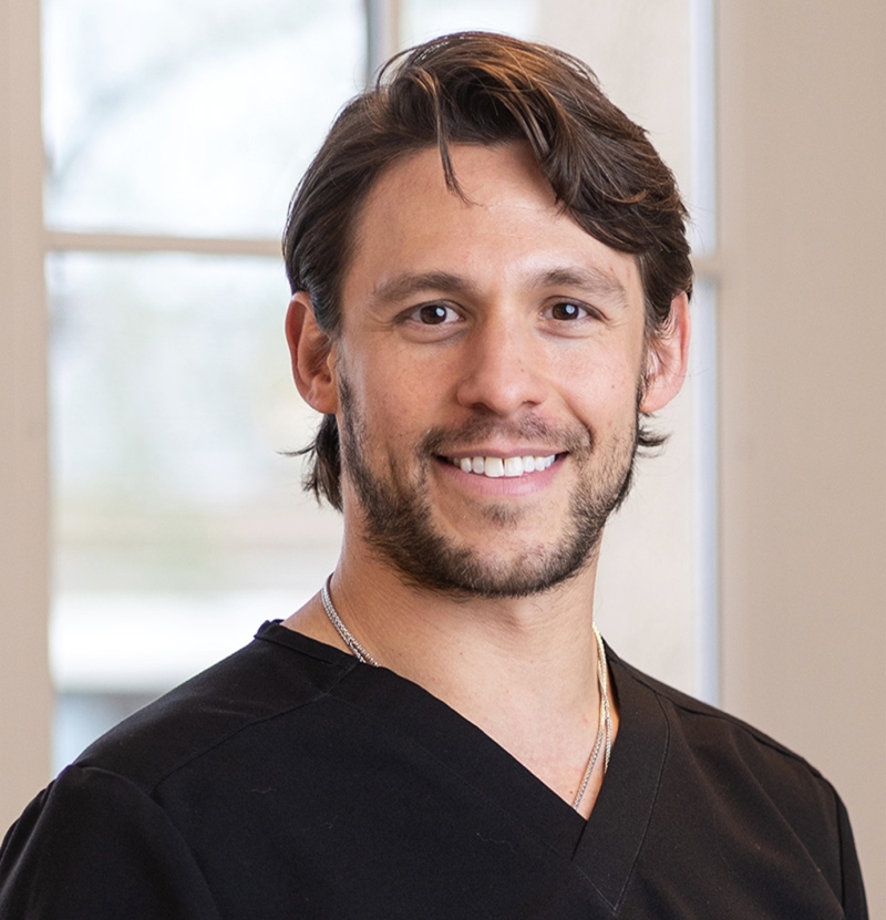 A man with medium-length brown hair and a beard, wearing a black V-neck top, smiles while standing indoors in front of large windows.