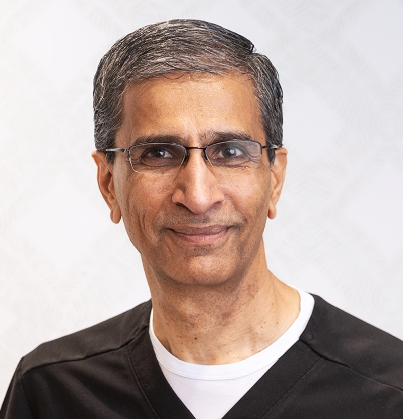 A middle-aged man with short gray hair and glasses, wearing a black and white shirt, smiles gently at the camera against a light, patterned background.