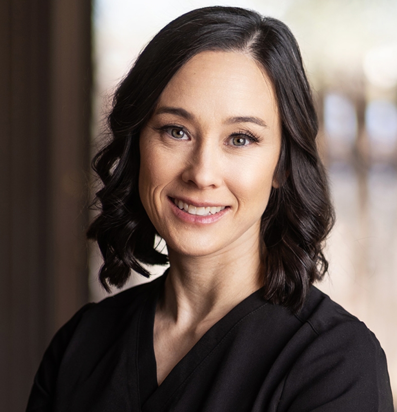 A woman with shoulder-length dark hair and light eyes smiles at the camera. She is wearing a black top and is standing indoors with a blurred background.