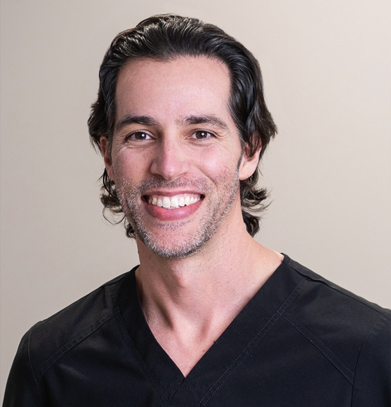 Smiling man with medium-length dark hair wearing a black V-neck scrub top, standing in front of a plain light background.