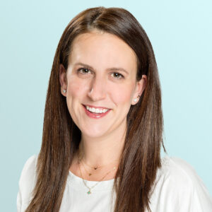 A woman with straight brown hair, wearing a white top and layered gold necklaces, smiles at the camera against a light blue background.