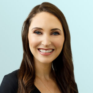 Smiling woman with long brown hair, wearing a black top, against a light blue background. She faces the camera, showing a friendly and approachable expression.