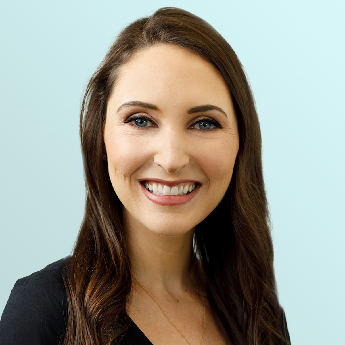 Smiling woman with long brown hair, wearing a black top, against a light blue background. She faces the camera, showing a friendly and approachable expression.