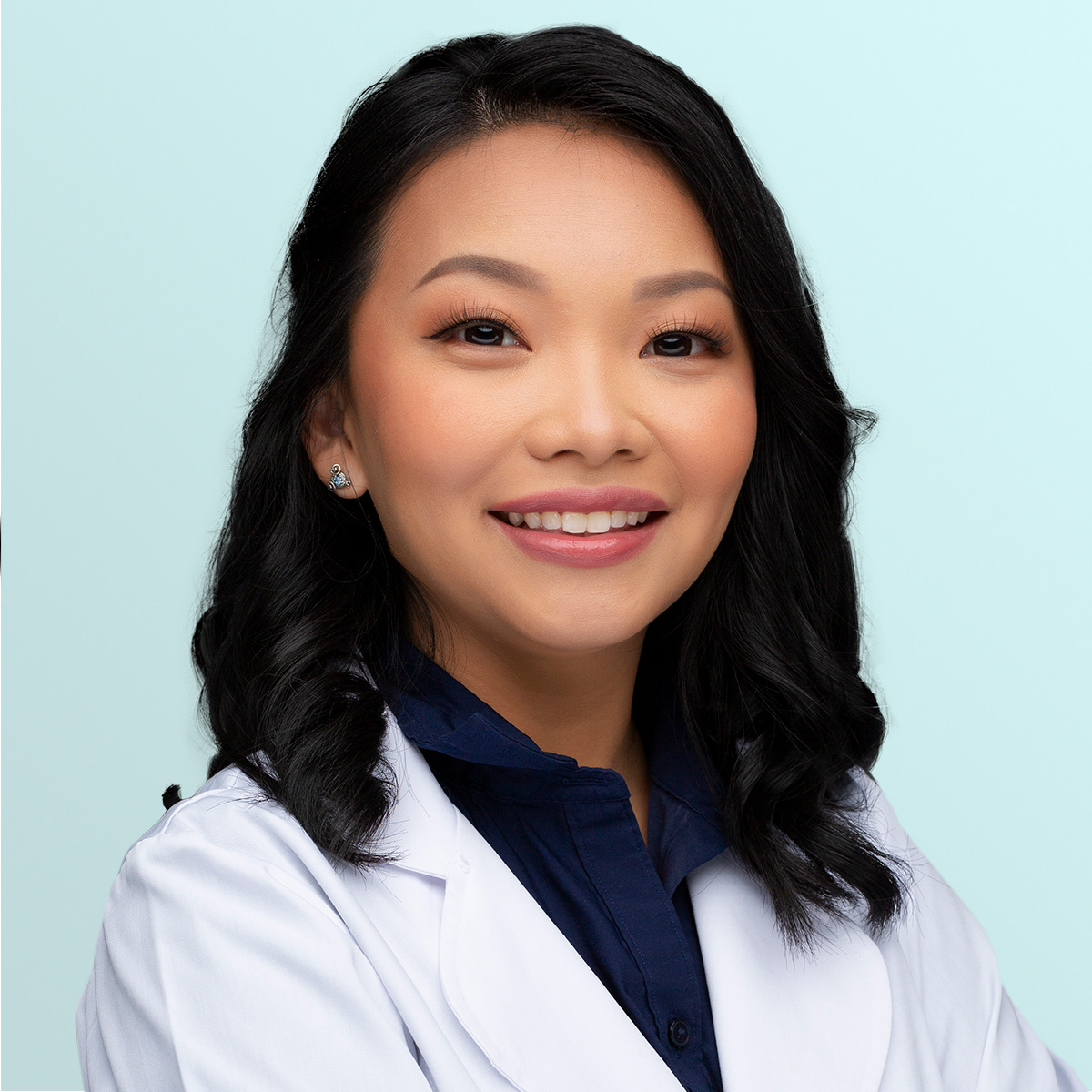 A woman with wavy black hair wearing a white lab coat and navy blouse smiles at the camera against a light blue background.