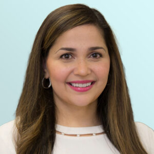 A woman with long brown hair, wearing a white top and hoop earrings, smiles at the camera against a light blue background.
