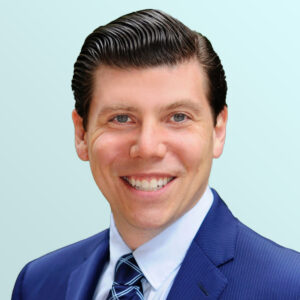 Smiling man with neatly combed dark hair wearing a blue suit, white shirt, and patterned blue tie, posing against a light blue gradient background.