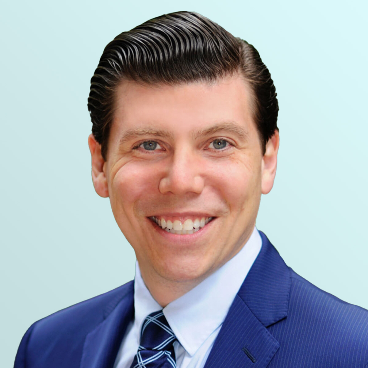 Smiling man with neatly combed dark hair wearing a blue suit, white shirt, and patterned blue tie, posing against a light blue gradient background.