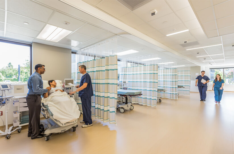 A patient in a hospital bed is attended by two medical professionals in a bright, modern hospital room with multiple curtained areas. Two other staff walk and talk in the background.