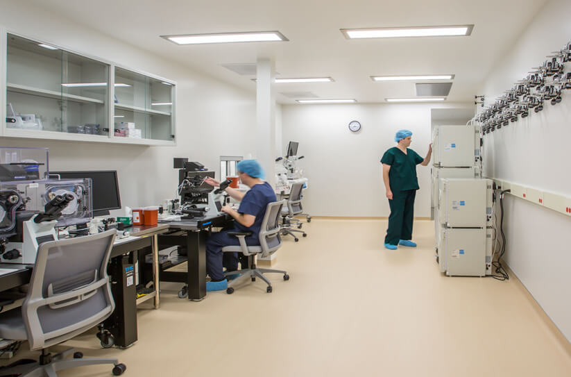 Two people in medical scrubs work in a bright, modern laboratory; one sits at a microscope while the other stands near large equipment along the wall. The room is clean and organized, with lab tools and computers visible.