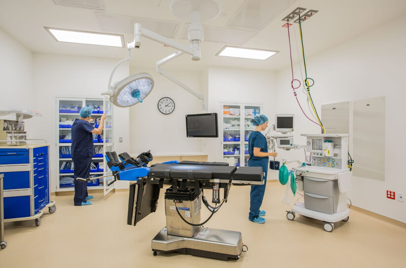 Two medical professionals in scrubs work in a bright, clean operating room with an operating table, medical equipment, storage cabinets, overhead lights, and a clock on the wall.