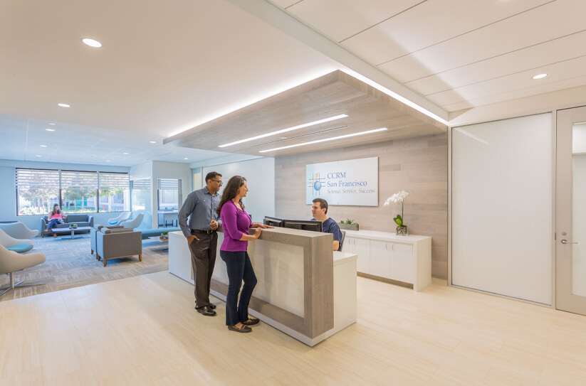 Two people stand at a modern clinic reception desk speaking with a receptionist. Behind the desk is a sign that reads CDRI San Francisco. The lobby has seating, large windows, and bright, clean decor.