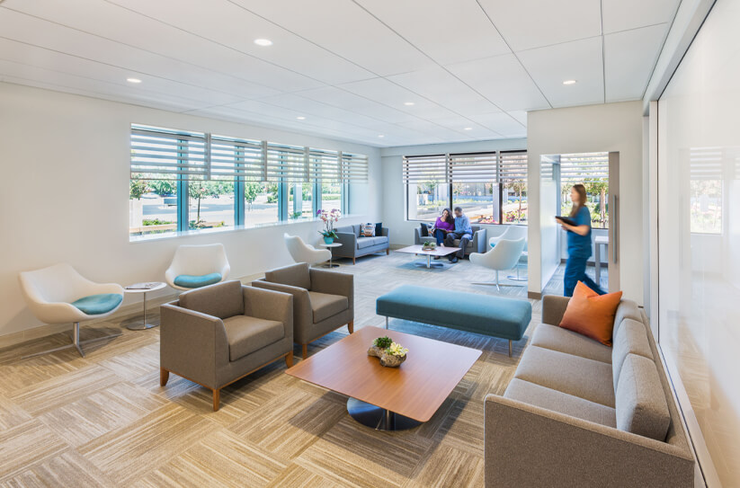 A modern waiting room with gray chairs, a blue ottoman, a wooden coffee table, large windows, and natural light. Two people sit and talk in the background while another person walks past.