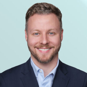 A man with short, light brown hair and a trimmed beard smiles at the camera. He is wearing a dark suit jacket over a light blue collared shirt, posed against a light blue background.