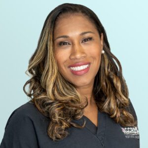 A woman with long, wavy, highlighted hair smiles at the camera. She is wearing pearl earrings and a dark scrub top with embroidered text, standing against a light blue background.