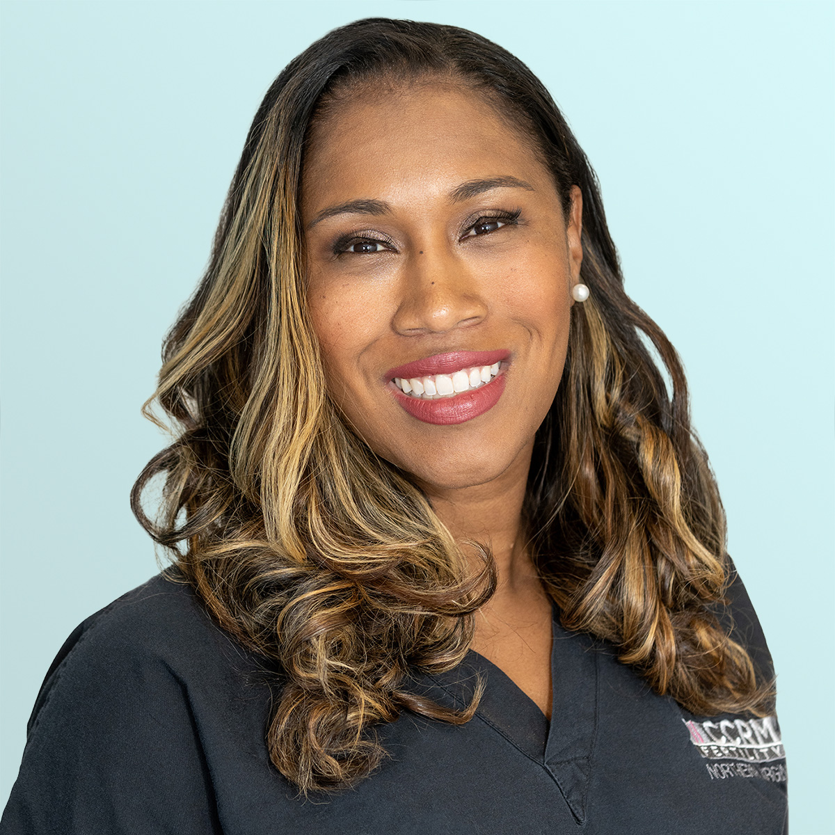 A woman with long, wavy, highlighted hair smiles at the camera. She is wearing pearl earrings and a dark scrub top with embroidered text, standing against a light blue background.