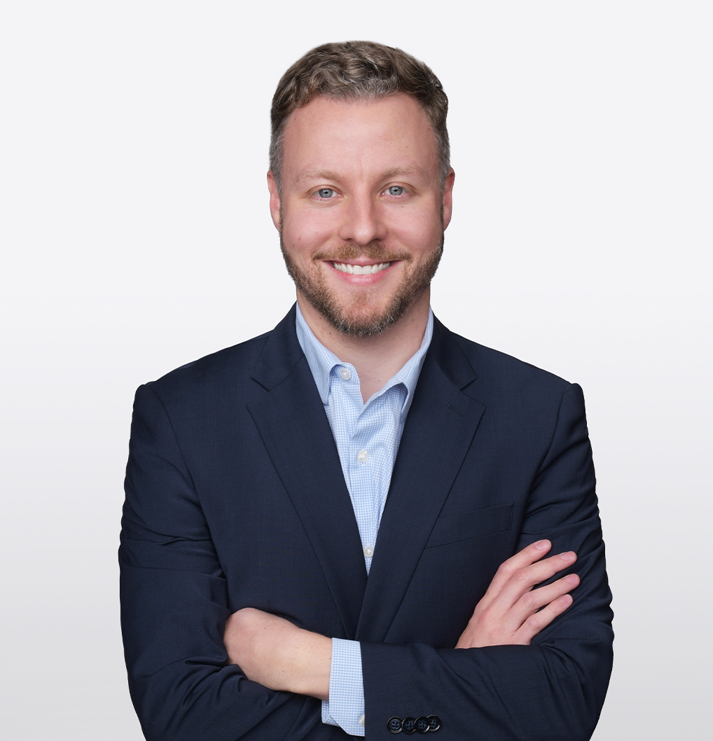 A man in a navy suit and light blue shirt stands smiling with arms crossed against a plain white background.