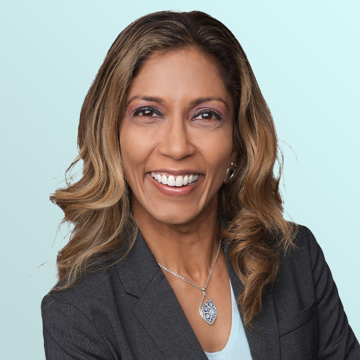 A woman with long wavy hair, wearing a dark blazer, light blue top, and a silver pendant necklace, smiles confidently against a light blue background.
