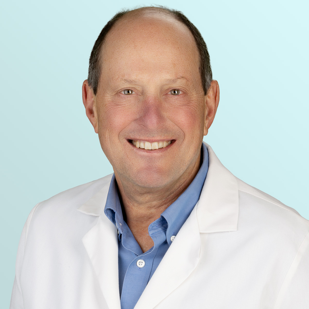 A smiling middle-aged man wearing a white lab coat over a light blue button-up shirt, standing against a light blue background.