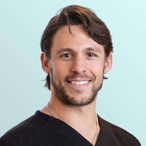 A smiling man with medium-length brown hair and a short beard, wearing a black V-neck shirt and a silver necklace, stands in front of a light blue background.