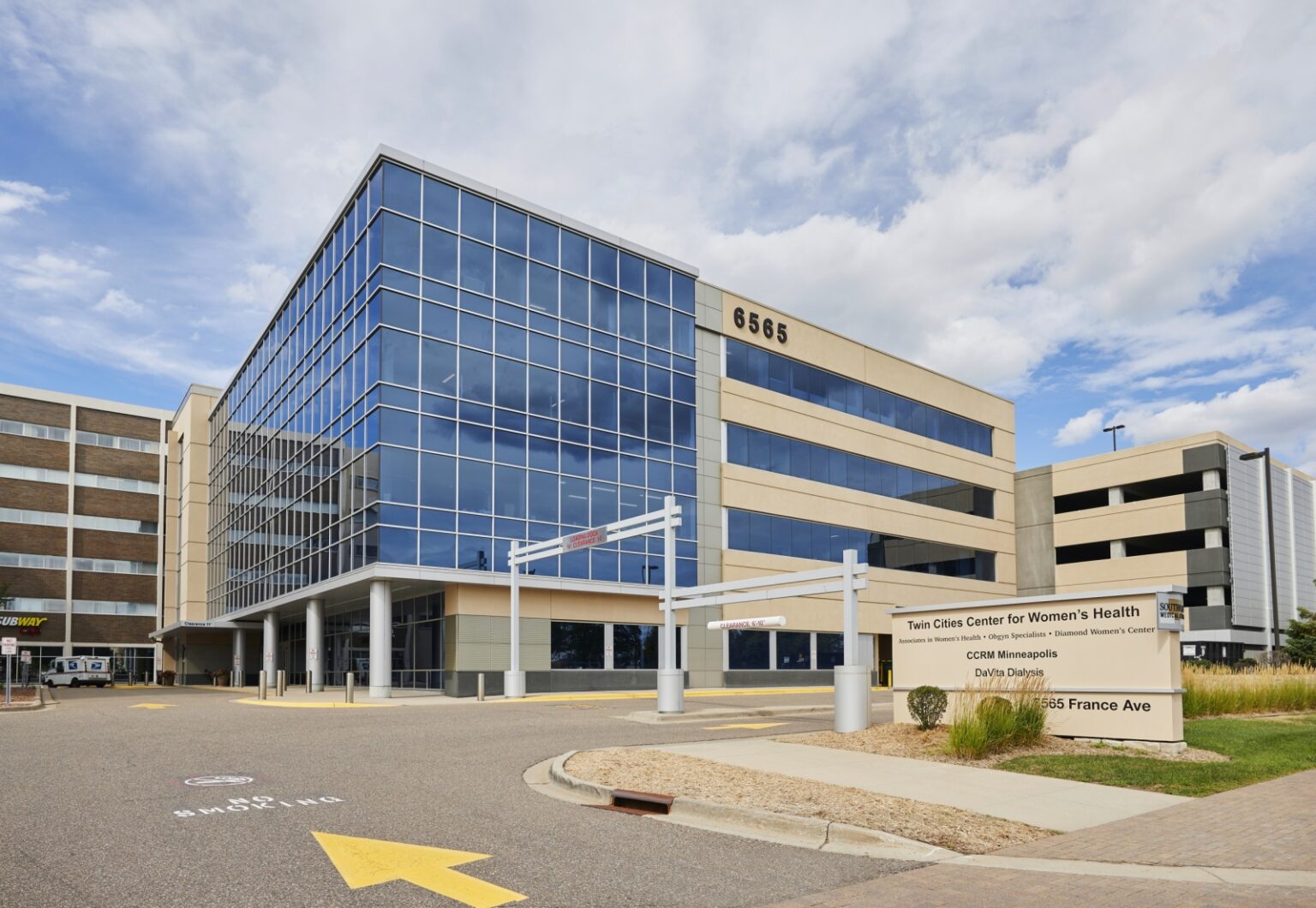 A modern glass and beige office building with the address 6565. A sign in front reads Twin Cities Center for Women’s Health and lists clinics and medical offices. The sky is partly cloudy.