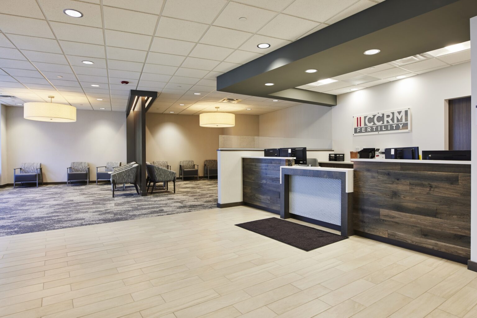 Modern medical office lobby with a front desk, CCRM Fertility sign on the wall, and a seating area with chairs and soft lighting in the background. The space has neutral tones and clean, contemporary design.