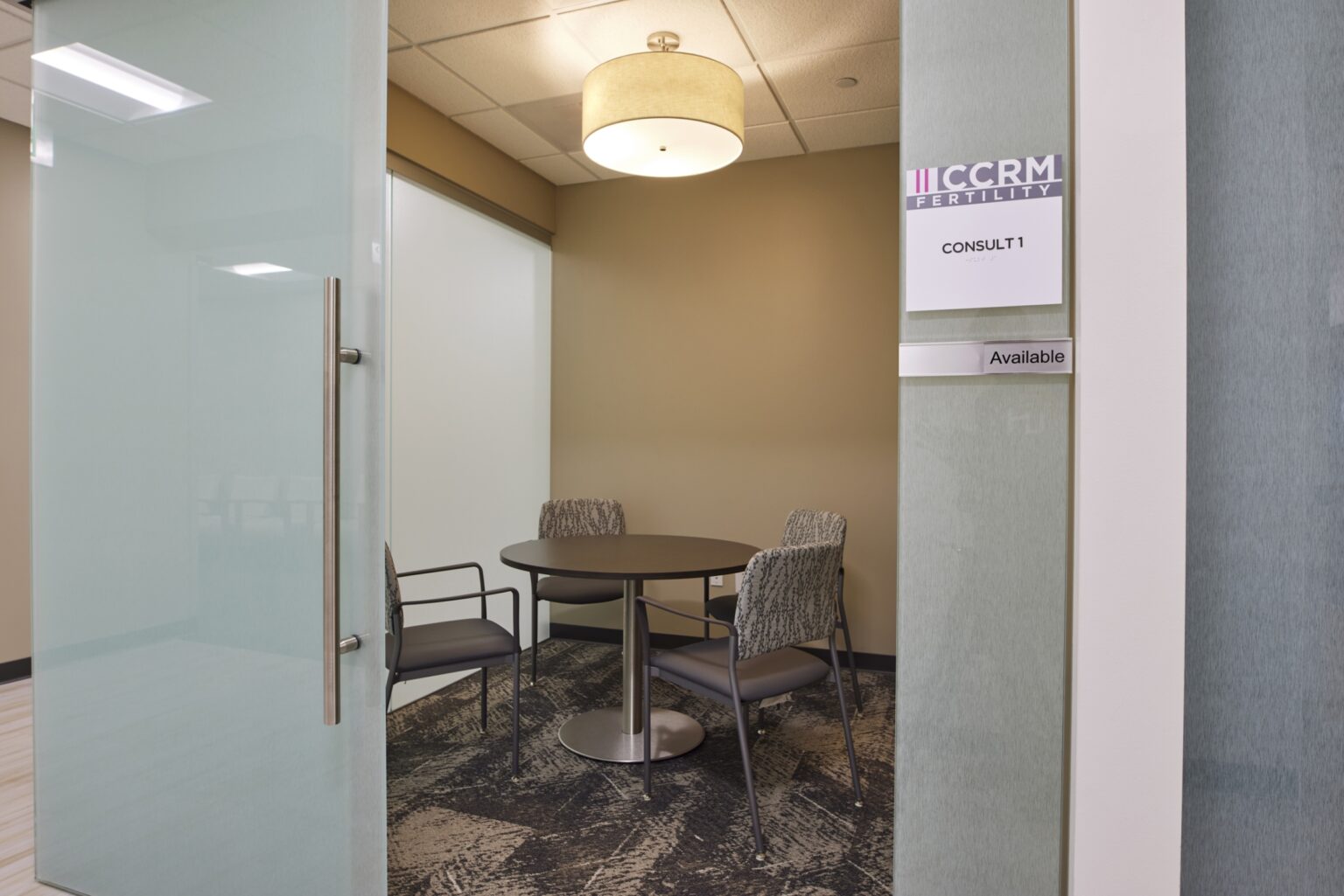 A small meeting room labeled Consult 1 with a round table, four chairs, and a ceiling light. The door is open, and a sign reads Available. The walls are beige and the floor has a patterned carpet.
