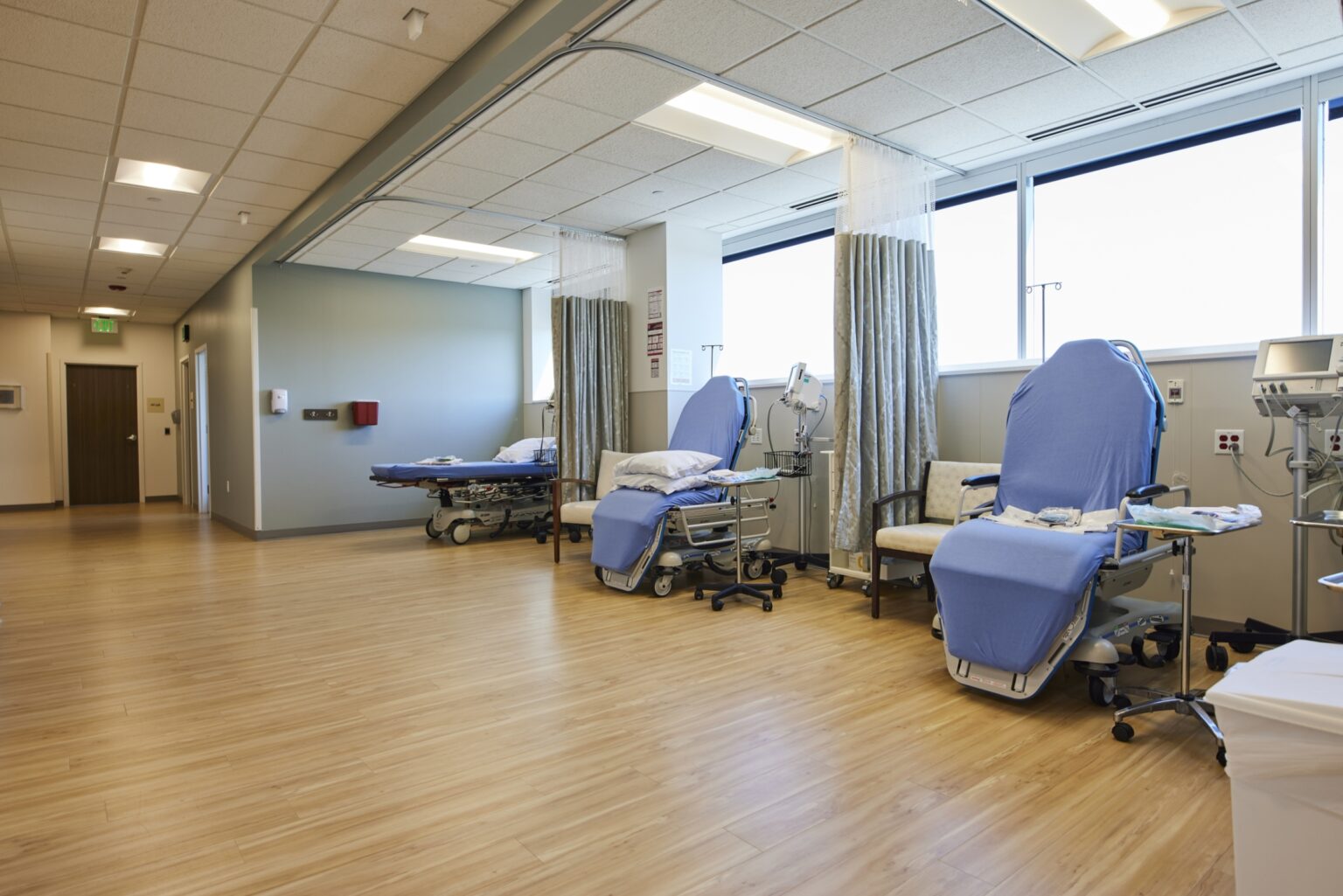 A bright, empty hospital room features several beds and chairs covered in blue sheets, separated by curtains, with medical equipment nearby and large windows letting in natural light.
