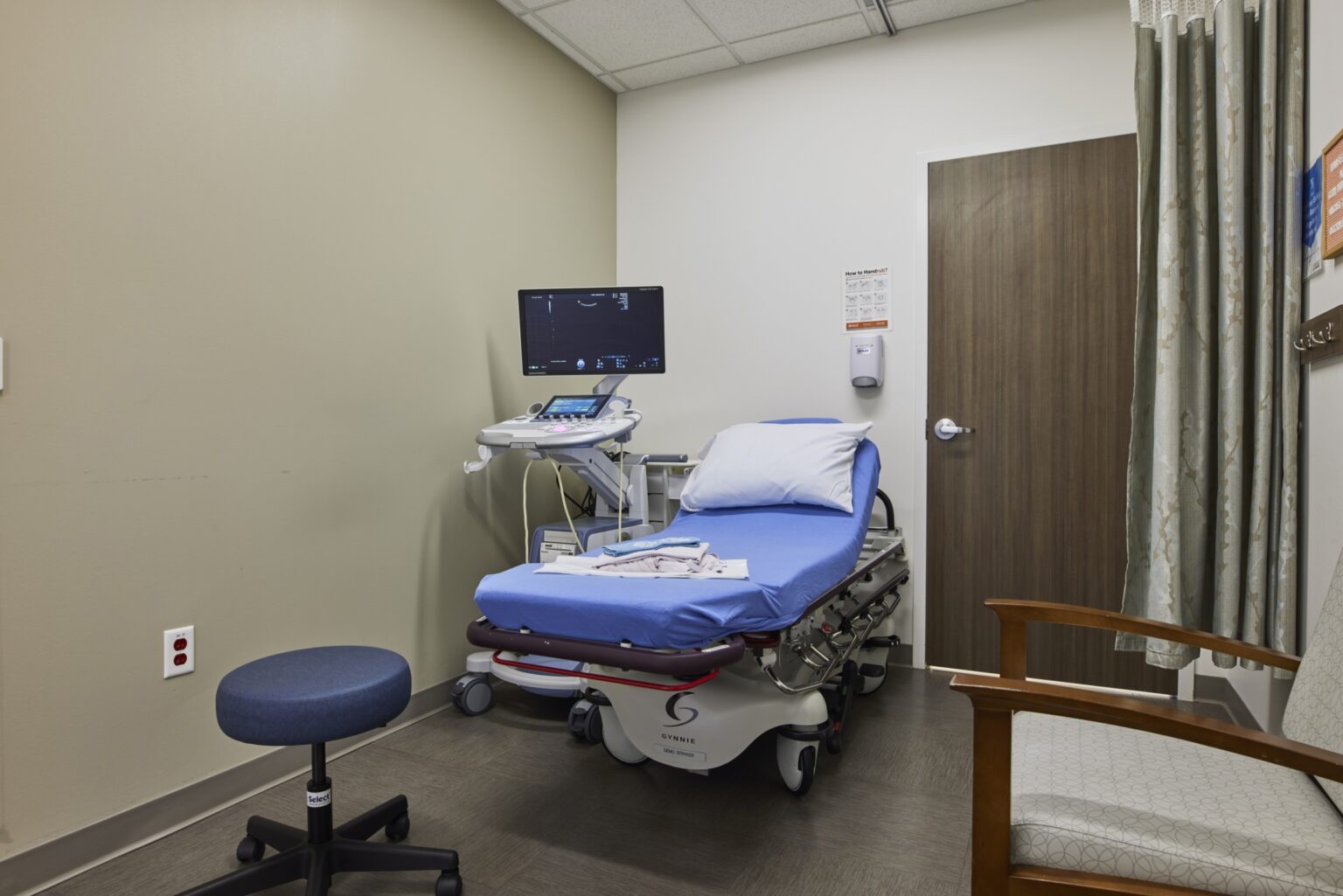 A small medical examination room with an exam bed, ultrasound machine, monitor, blue stool, wooden chair, and a closed wooden door. The bed is covered with a blue sheet and a pillow. The walls and floor are plain.