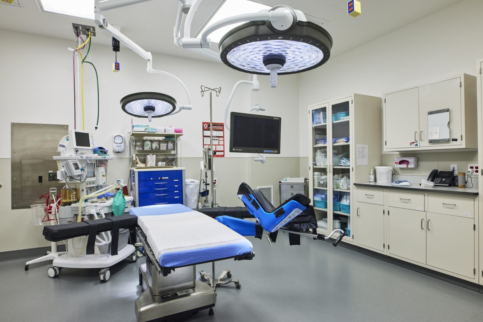 A modern operating room with surgical lights, an operating table, medical monitors, equipment carts, and storage cabinets, all clean and organized, ready for use.