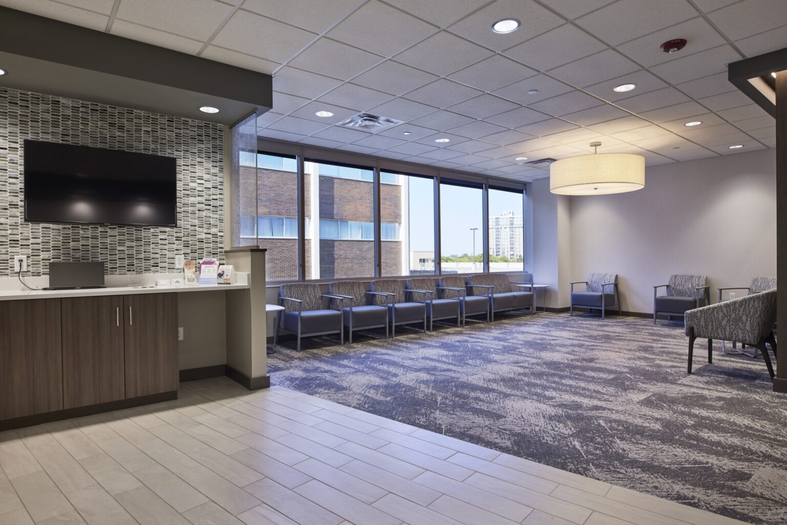 Modern waiting room with large windows, rows of blue chairs, patterned carpet, a wall-mounted TV, and a refreshment counter. The space is brightly lit with ceiling lights and a large round hanging light fixture.