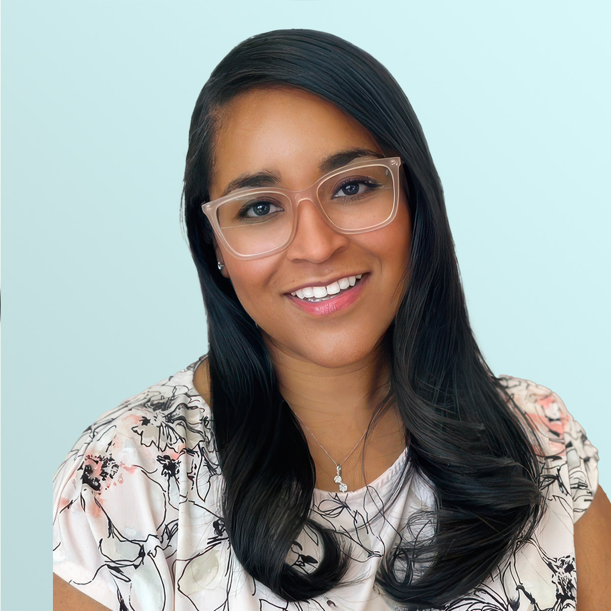 A woman with long, dark hair and clear-framed glasses smiles at the camera. She is wearing a light-colored, floral top and a necklace, with a pale blue background behind her.