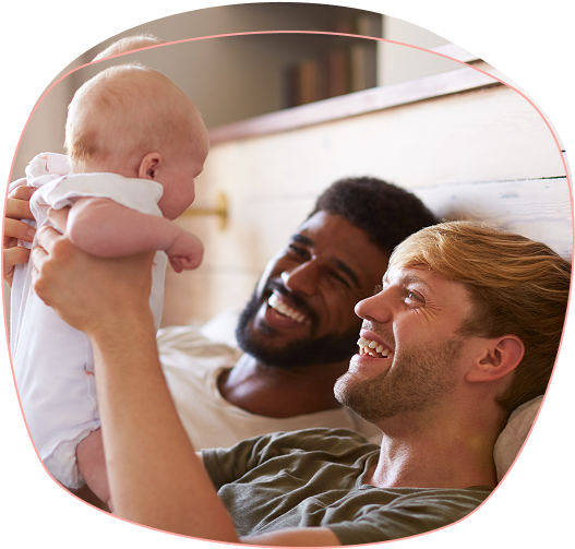 Two men lying on a bed smiling and holding a baby, engaging warmly with the child. The scene conveys a sense of happiness and family bonding.