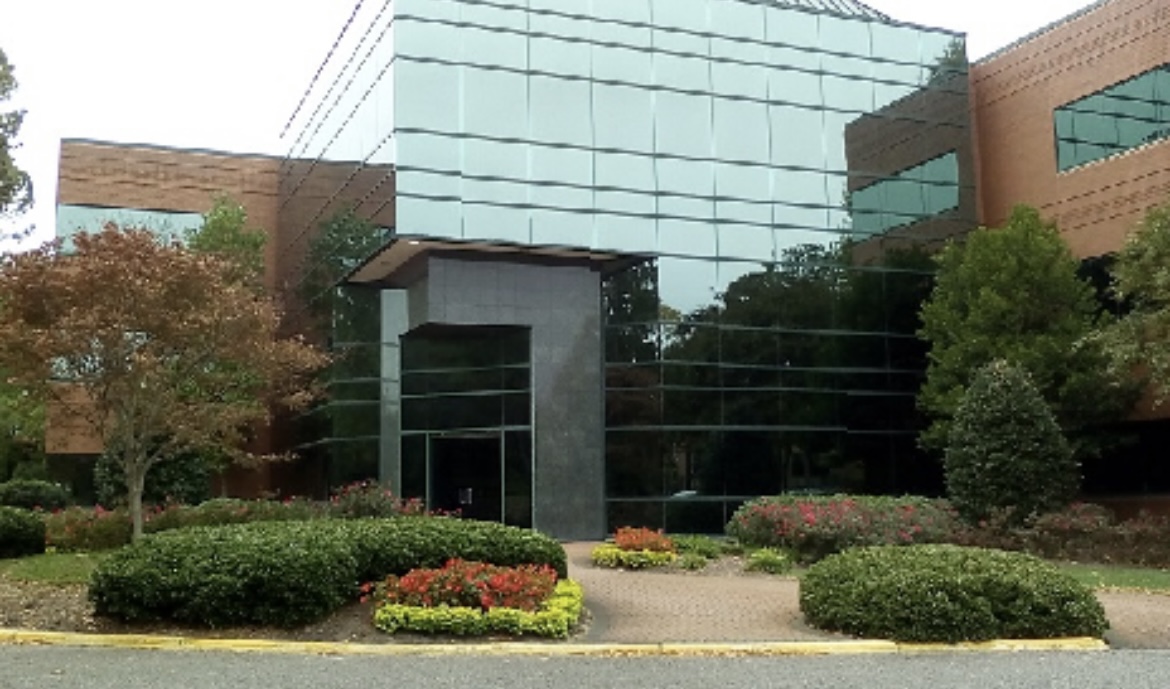 Modern office building with large glass windows and brown brick exterior, surrounded by neatly landscaped bushes, flowers, and small trees.