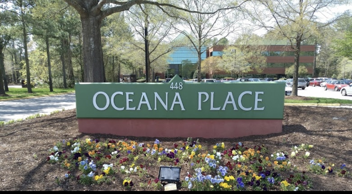 Large green sign reading OCEANA PLACE 448 stands in front of a bed of colorful flowers, with trees and a modern brick-and-glass office building in the background. Several cars are parked in the lot.