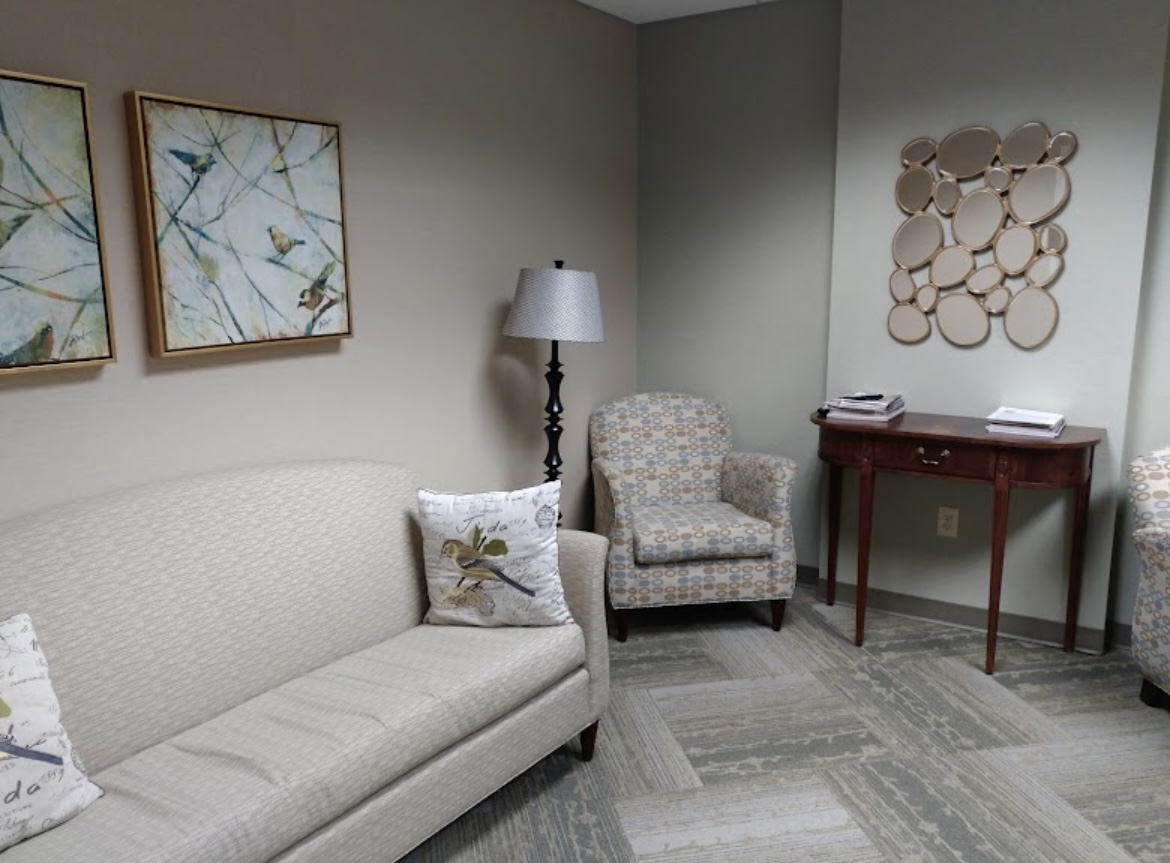 A waiting room with a beige sofa, patterned armchair, small wooden table with magazines, standing lamp, and bird-themed wall art. The room has neutral colors and a soft, modern décor.