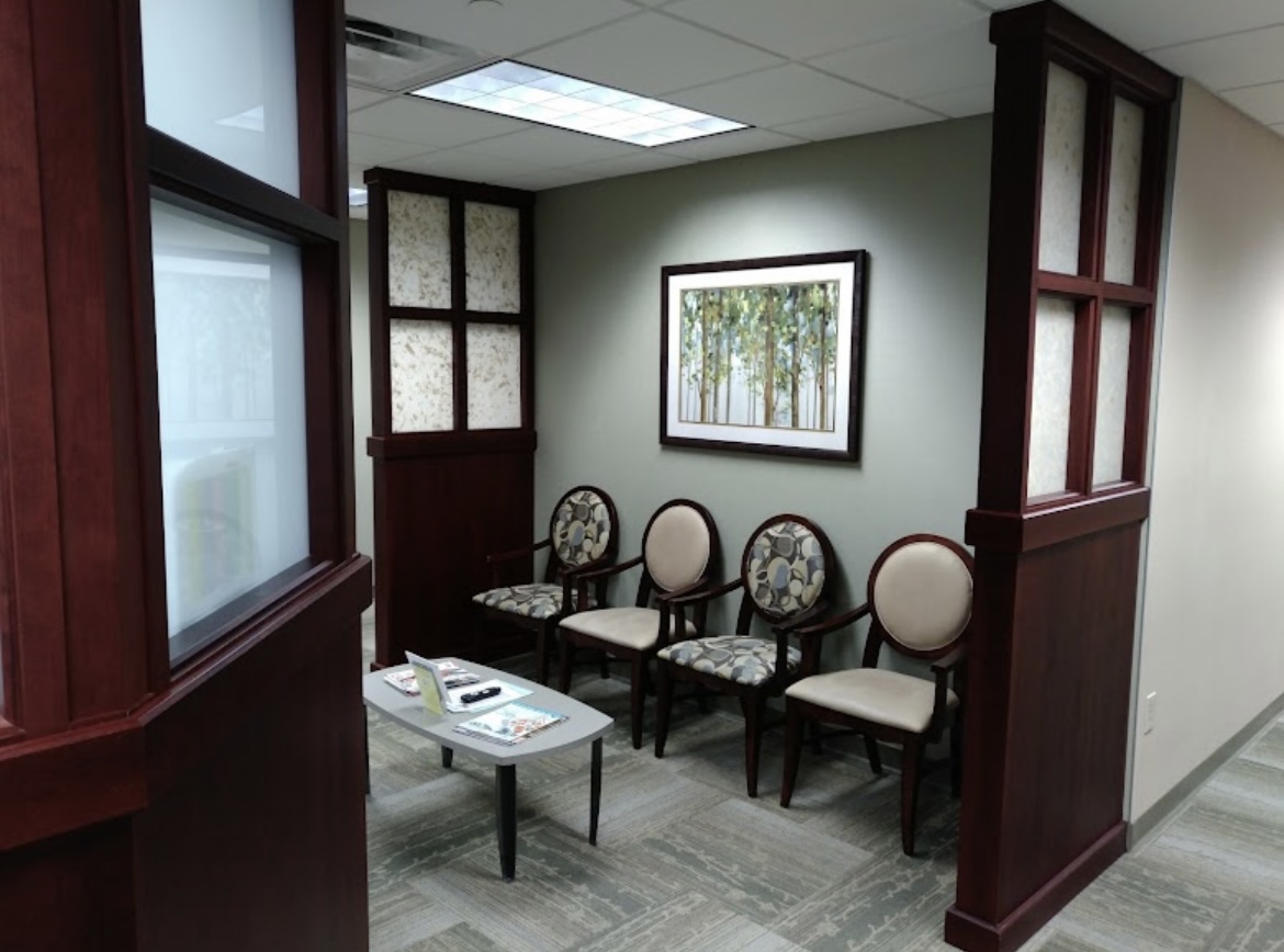 A small waiting area with four cushioned chairs, a low table with magazines, and a framed painting of trees on the wall. The space is enclosed with dark wood panels and has a neutral color scheme.