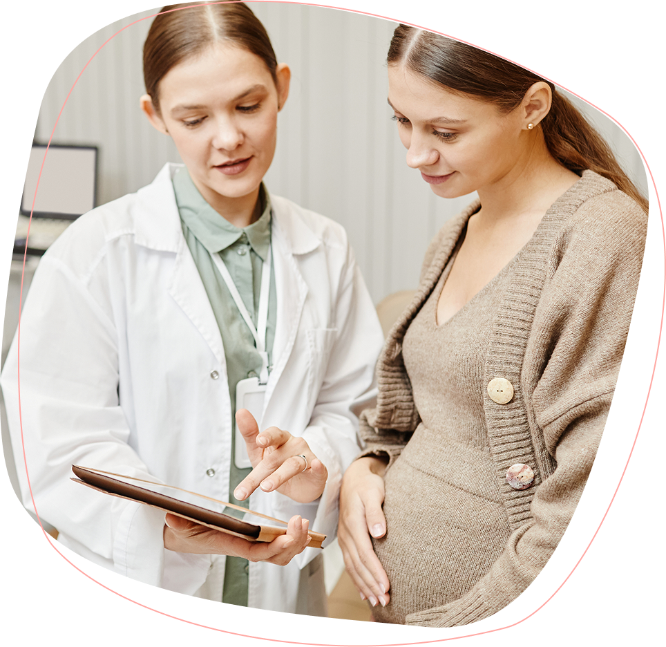 A doctor in a white coat shows information on a tablet to a pregnant woman, who is gently touching her belly. Both are standing and appear focused on the screen.