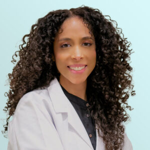 A woman with long, curly hair wears a white lab coat and a black shirt, smiling at the camera against a light blue background.