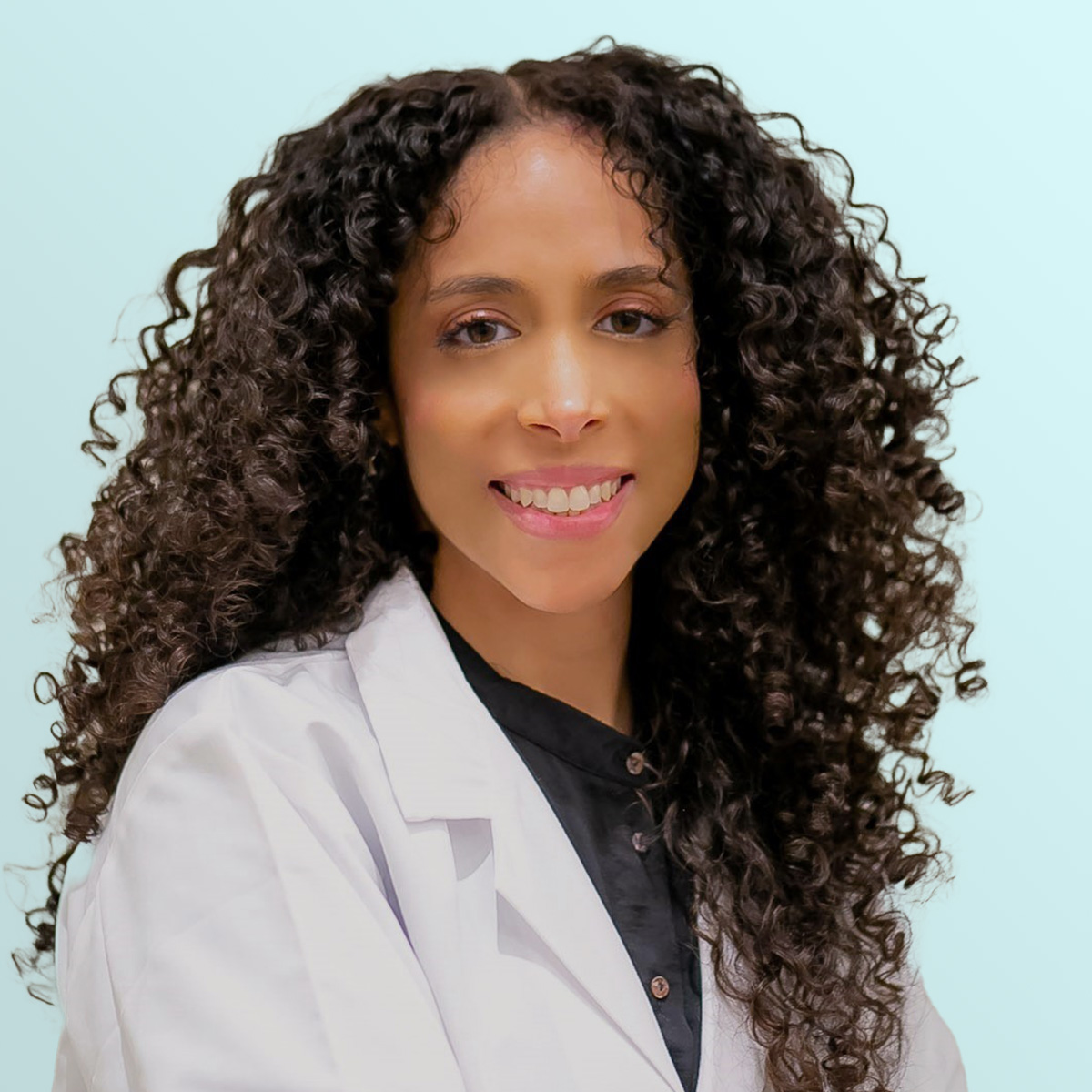 A woman with long, curly hair wears a white lab coat and a black shirt, smiling at the camera against a light blue background.