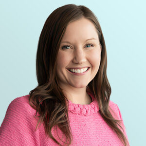 A woman with long brown hair, wearing a bright pink sweater, smiles at the camera against a light blue background.