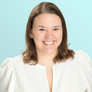A woman with shoulder-length brown hair smiles at the camera. She is wearing a textured white top and is posed in front of a light blue background.