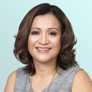 Smiling woman with shoulder-length wavy brown hair, wearing a gray sleeveless top and a gold necklace, posed in front of a light blue background.