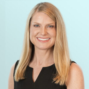 Smiling woman with straight blonde hair wearing a sleeveless black dress, standing against a light blue background.