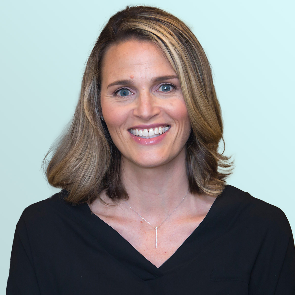 A woman with shoulder-length light brown hair and a side part, wearing a black V-neck top and a simple necklace, smiles in front of a light blue background.