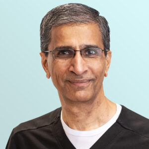 A middle-aged man with short gray hair and glasses, wearing a black shirt over a white undershirt, smiles slightly at the camera against a light blue background.
