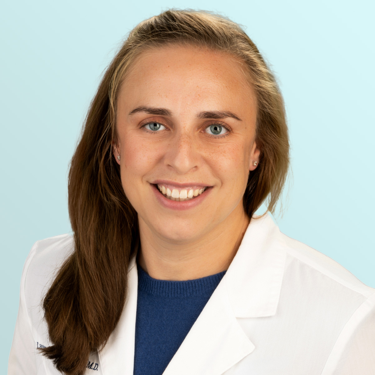 A woman with light brown hair and blue eyes, wearing a white lab coat over a blue top, smiles at the camera in front of a light blue background.