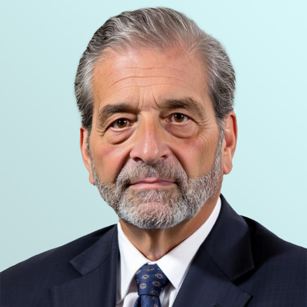 A mature man with gray hair and beard, wearing a navy suit, white shirt, and patterned tie, poses against a light blue background.