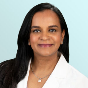 A woman with long dark hair, wearing a white coat, earrings, and a necklace, smiles at the camera against a light blue background.