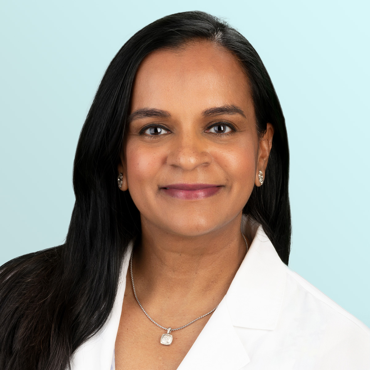 A woman with long dark hair, wearing a white coat, earrings, and a necklace, smiles at the camera against a light blue background.